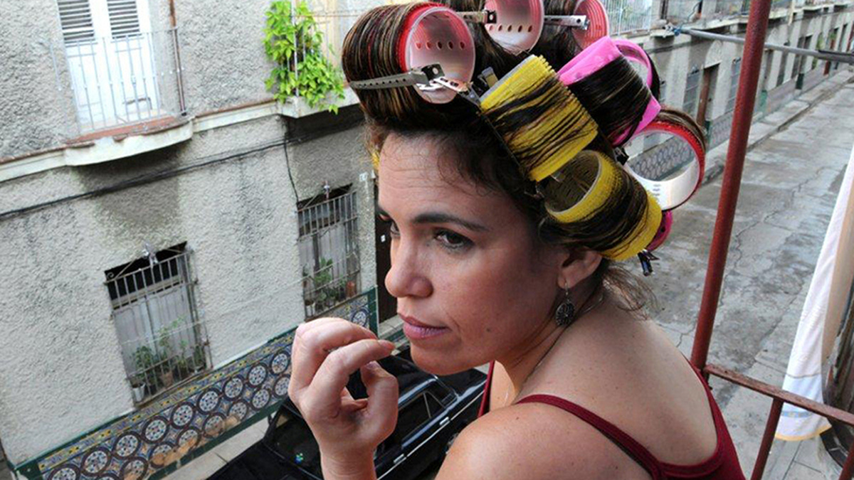 A woman with yellow and pink curlers in her hair looks out onto a narrow street from a balcony.
