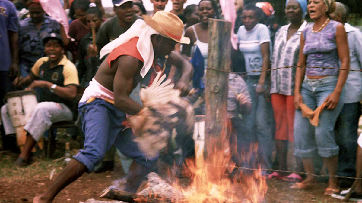 A man holds a chicken by a fire surrounded by a crowd and a drummer.