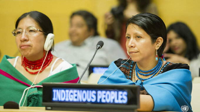 Two women in Indigenous clothing sitting at a wooden desk with headphones and sign that reads 'Indigenous Peoples'