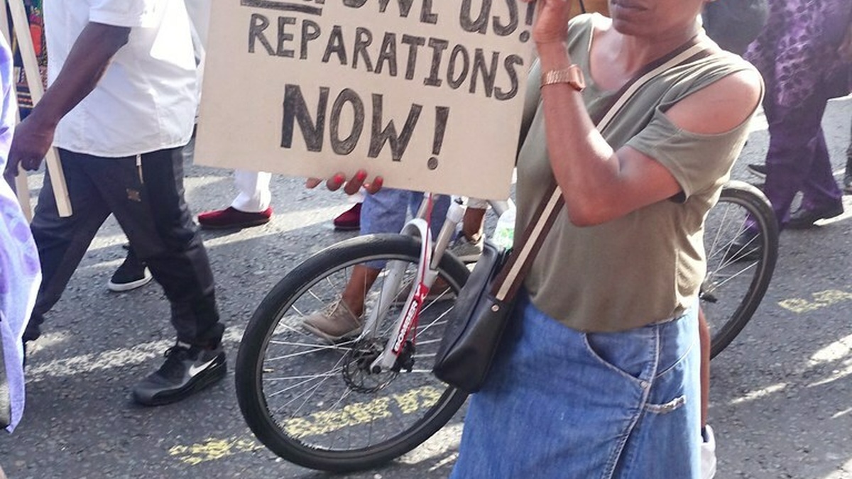 Women with placard that reads "You sold us! You stole us! You owe us! Reparations now" standing in front of a crowd.