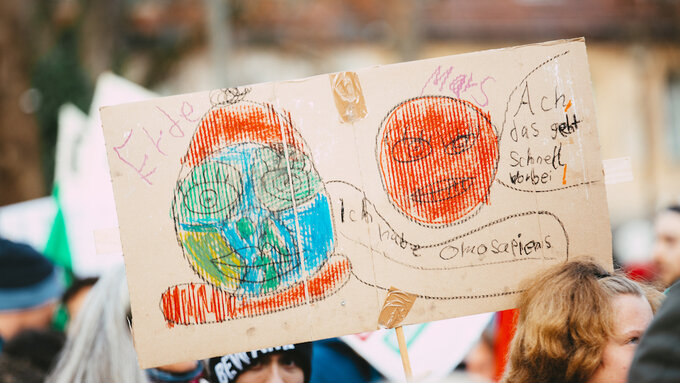 Cardboard placard with child's drawing of Earth and Mars held above blurred crowd.