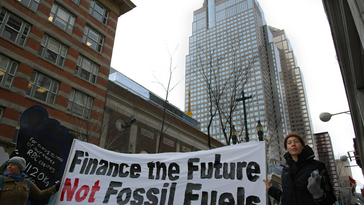 Two people holding up a banner that reads "Finance the Future. Not Fossil Fuels" in front of 2 high rise buildings.