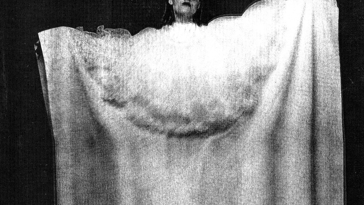 A black and white image of a person wearing a large white cloak, arms outstretched.