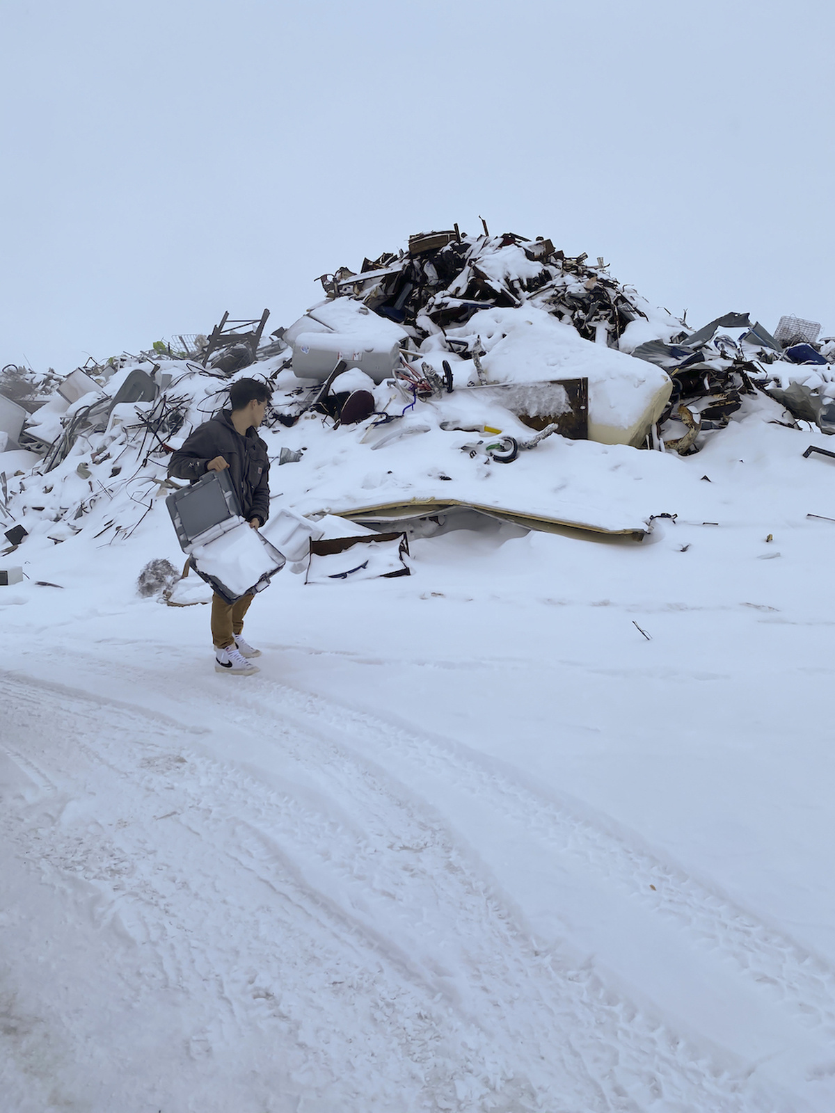 A person throws a suitcase on to a pile of discarded furniture and other rubbish. The ground and pile is covered in snow