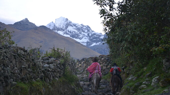 A landscape featuring mountains in the distance. Two people on horseback ride towards the mountains.