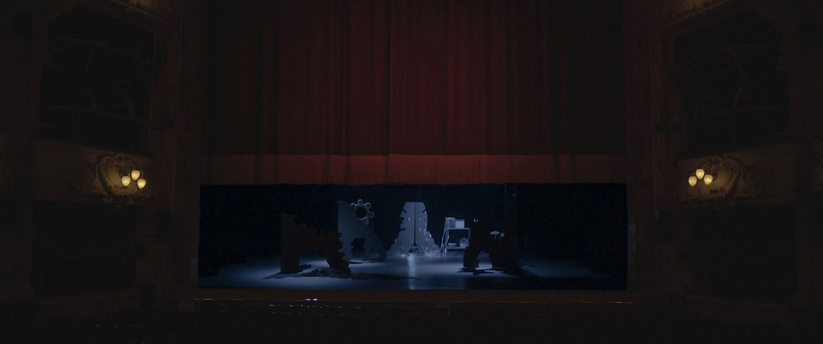 From an audience perspective looking onto a darkly lit ornate Victorian theatre stage with luscious red velvet curtains.