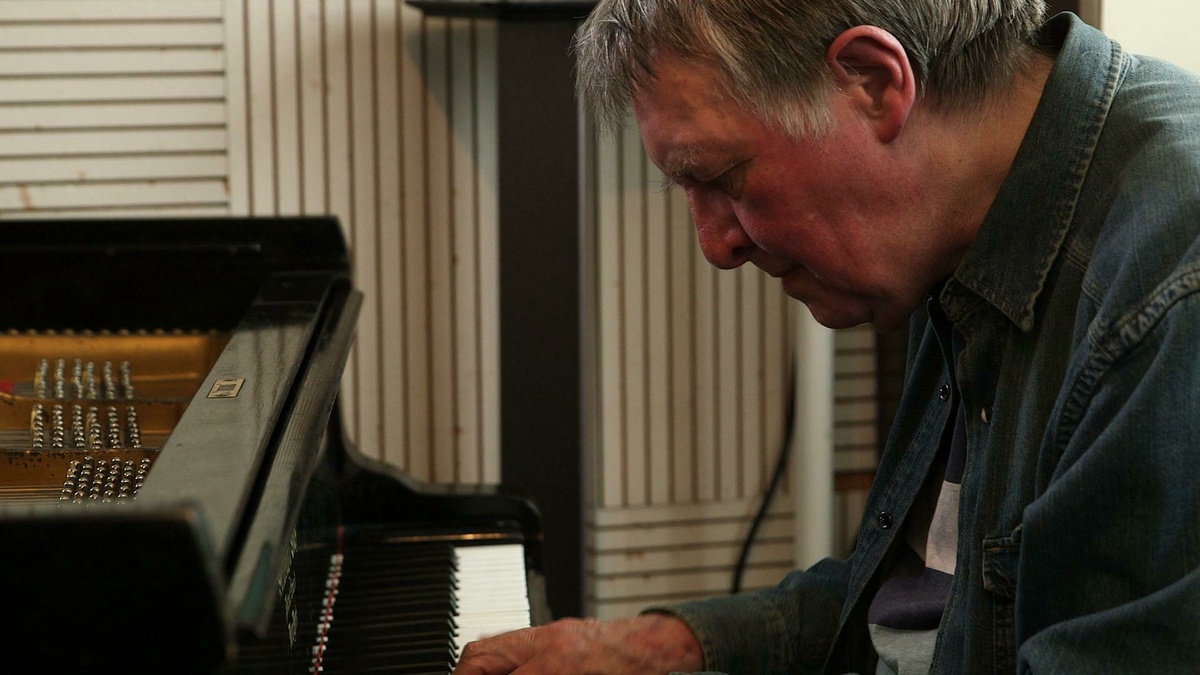 A photograph of Howard Riley, he leans forward at a piano.