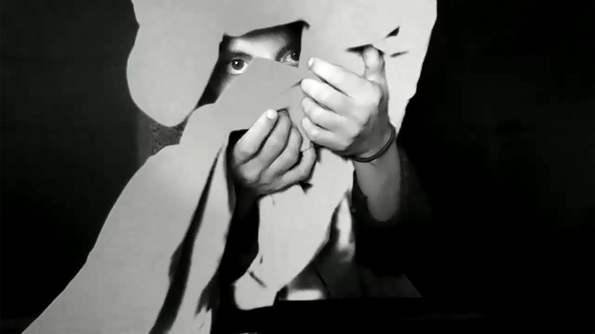A black and white still, a person peeks through a cut out abstract shape.