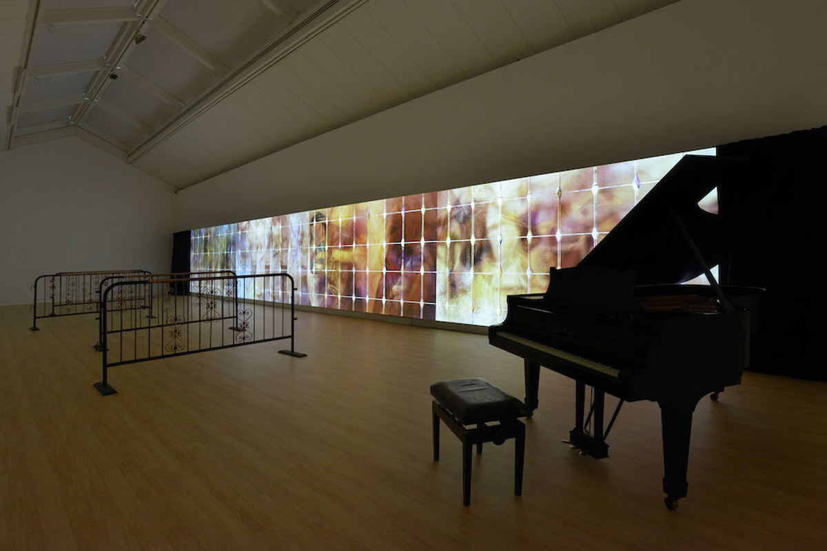 A gallery space with a colourful projection on one wall. There is a grand piano within the space.