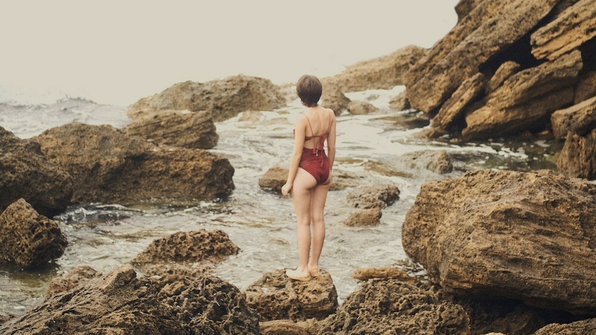 A white woman with short hair and a red bathing suit is standing on sea rocks. Her back is turned towards the camera.