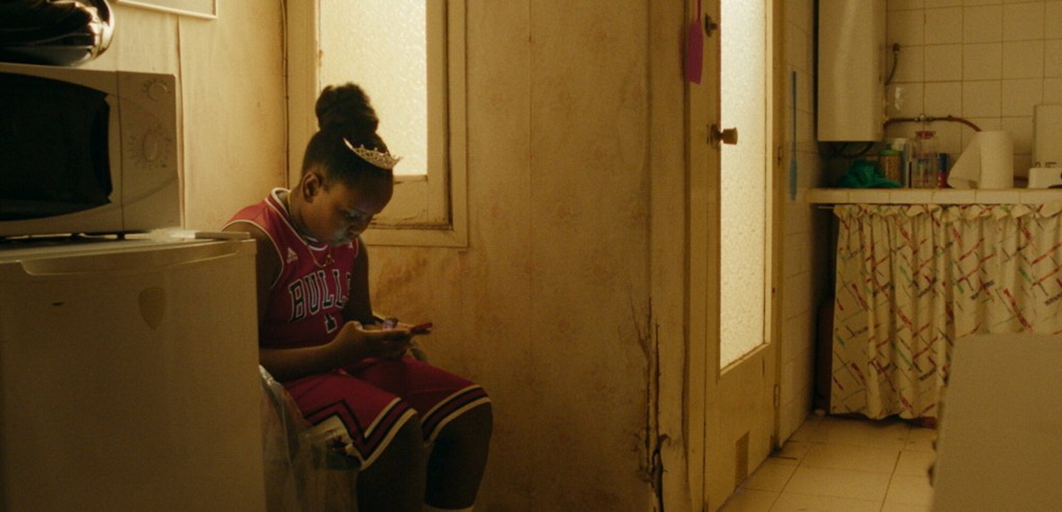 A young girl with black skin and dark, curly hair is sitting in a worn-down kitchen, looking down at a smartphone.