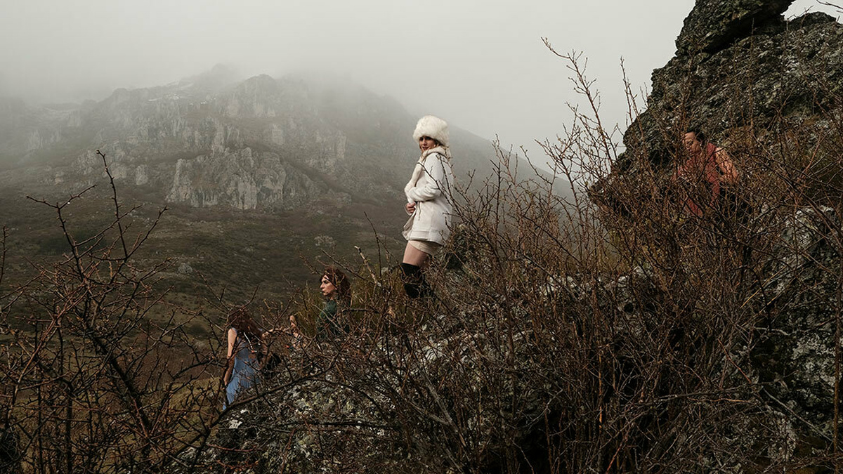 A group of trans women standing on a rock, surrounded by bushes. In the background, a mountain peak is covered by fog.