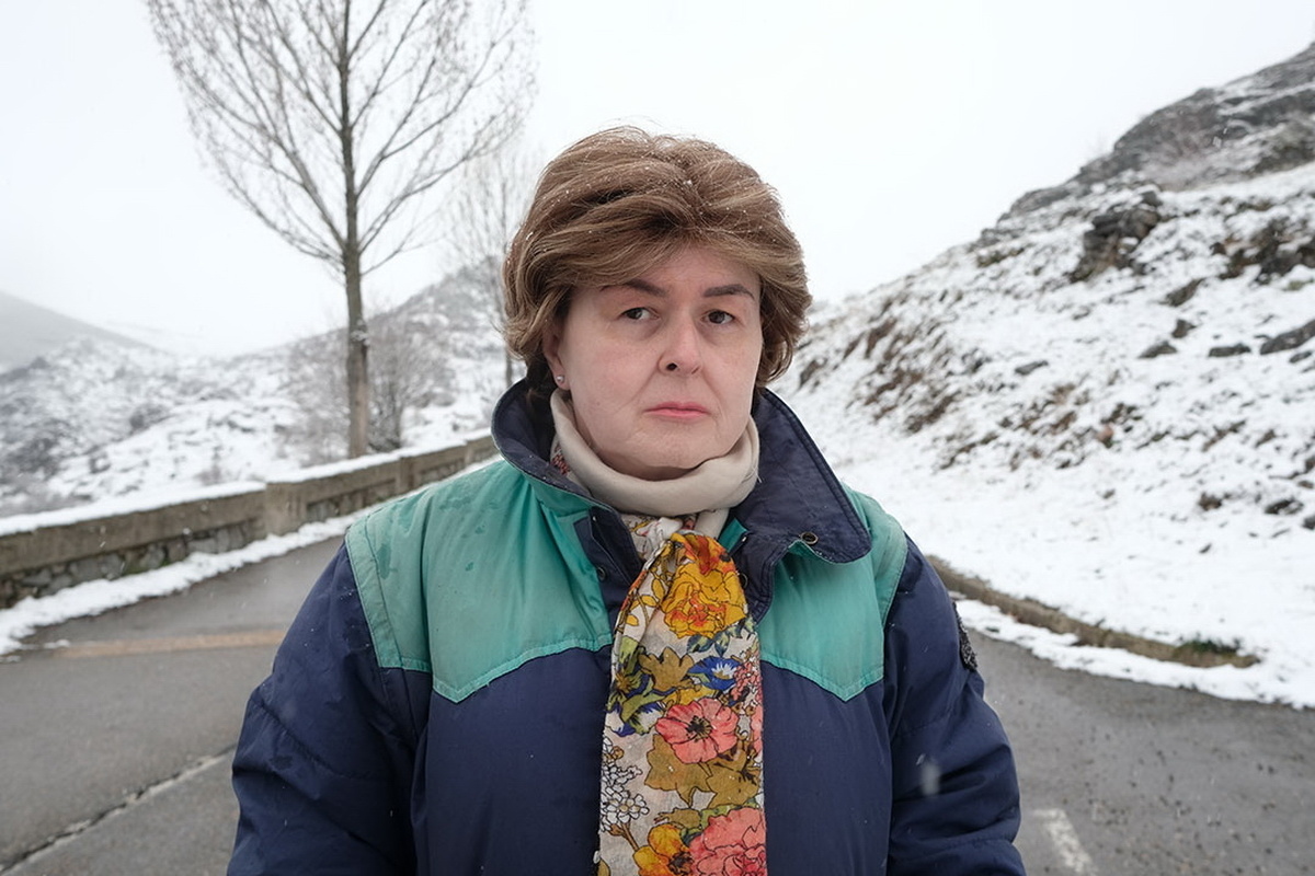 A trans woman with short, brown hair is standing on a mountain road, looking at the camera with a serious expression.