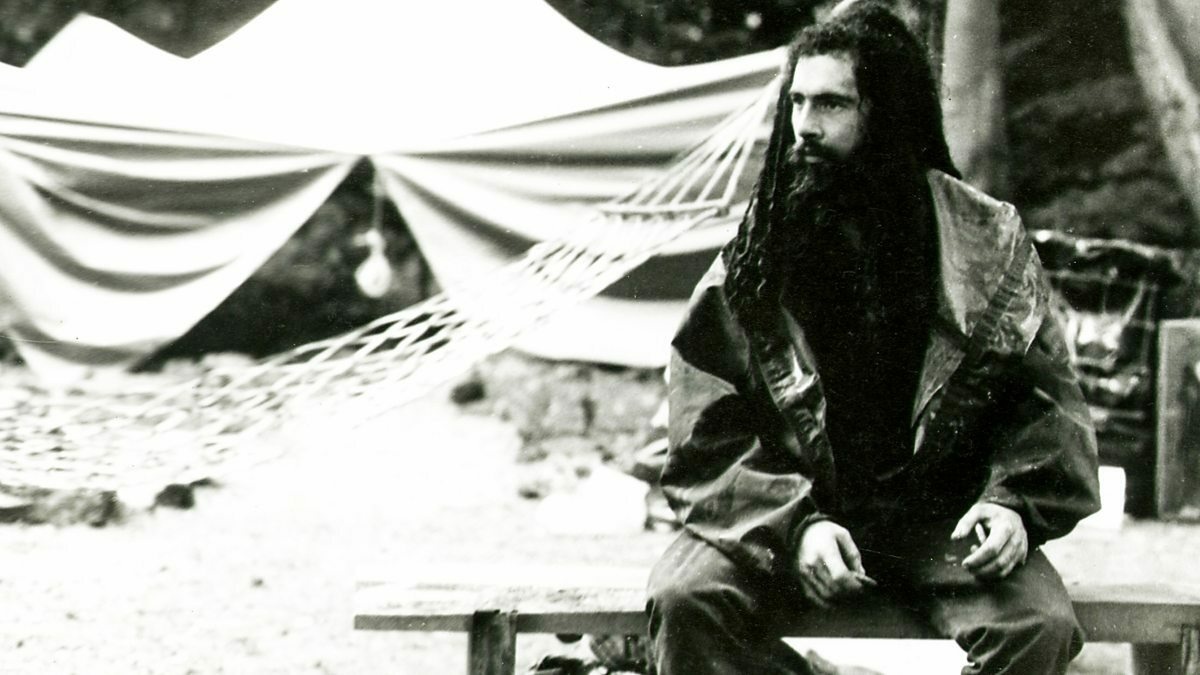 A man with dreadlocks and beard is sitting in woodland in front of a makeshift tent and hammock.