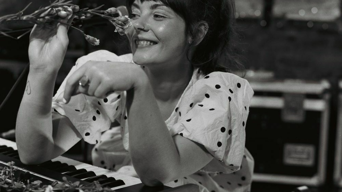 A black and white image, of a woman sitting at a keyboard, holding a flower up to her face and smiling.