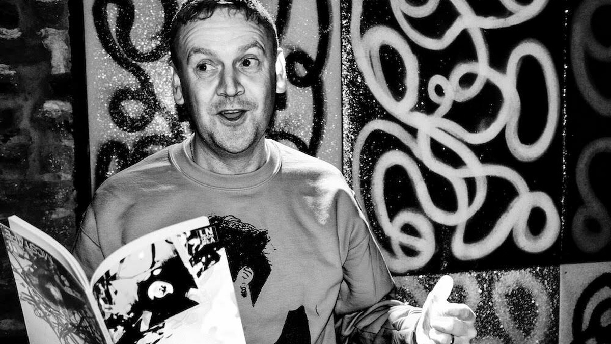 A man with short hair holding a book while performing poetry at an event. Behind him is a wall with graffiti.