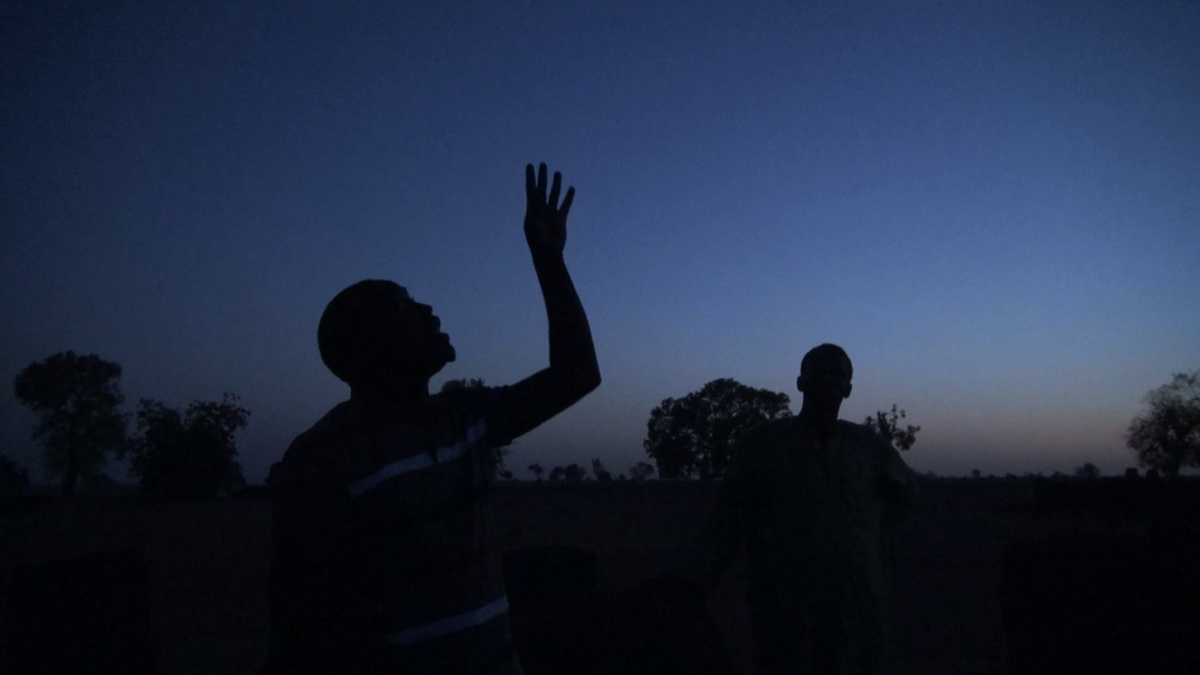 A video still, two people silhouetted against the sky at twilight. The figure on the left raises their hand to the sky.