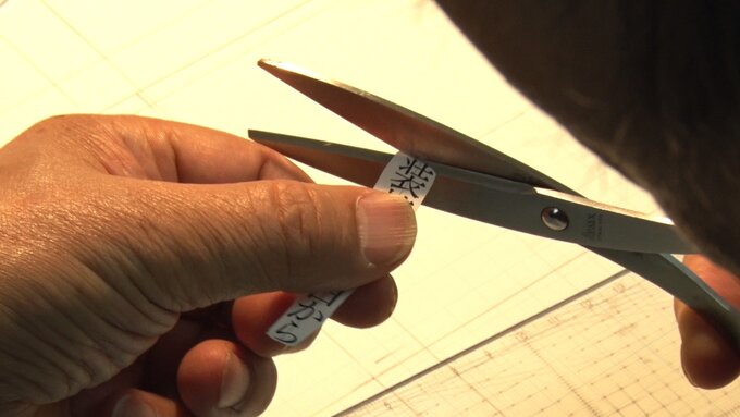 Hand holding text printed on paper being cut by a pair of scissors, with graphic paper in the background.