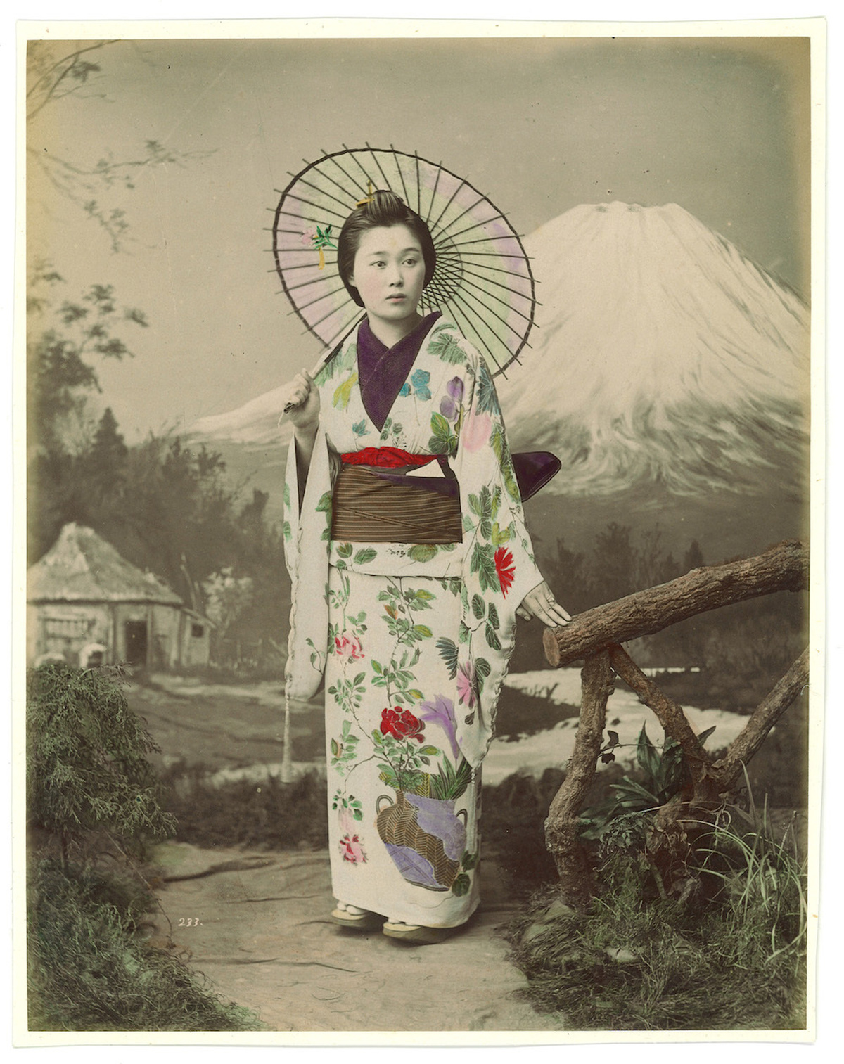 A woman in traditional Japanese attire posing for a photograph with Mount Fuji and a hut in the background.