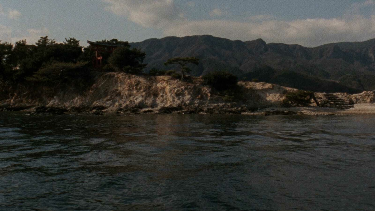 Island with Shinto shrine emerging from green foliage, hilly terrain in the background and sea water in the foreground.