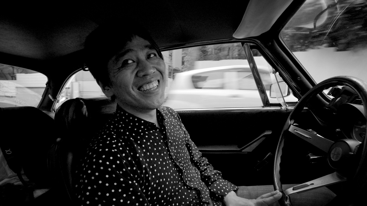 Monochromatic image of a man sitting behind the wheel of a car, grinning, with traffic in the background.