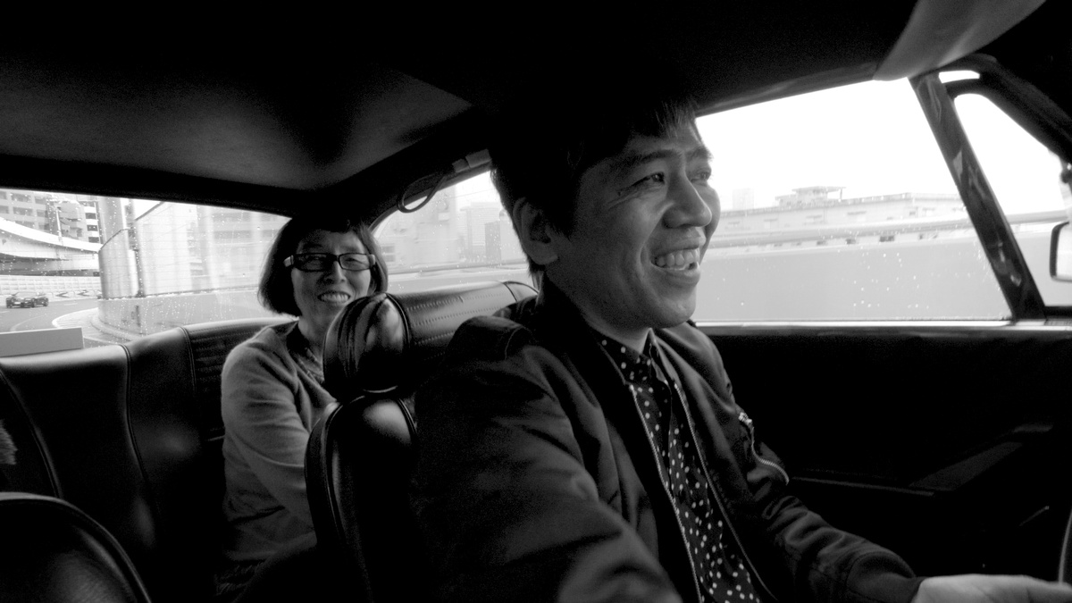 Monochromatic image of a smiling man driving a car with a woman behind him smiling, a motorway in the background.