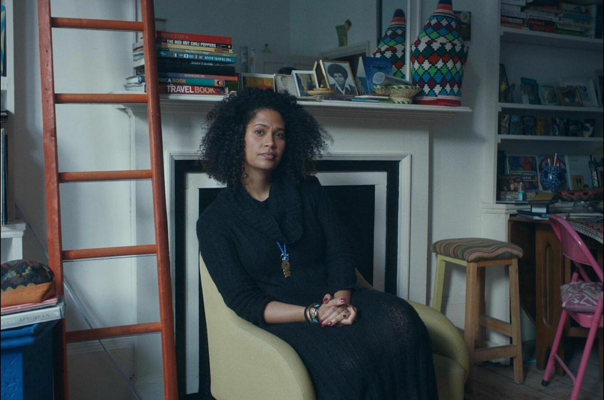 A black woman in a black dress sits in an arm chair looking into the camera. There are books on the shelf behind her.