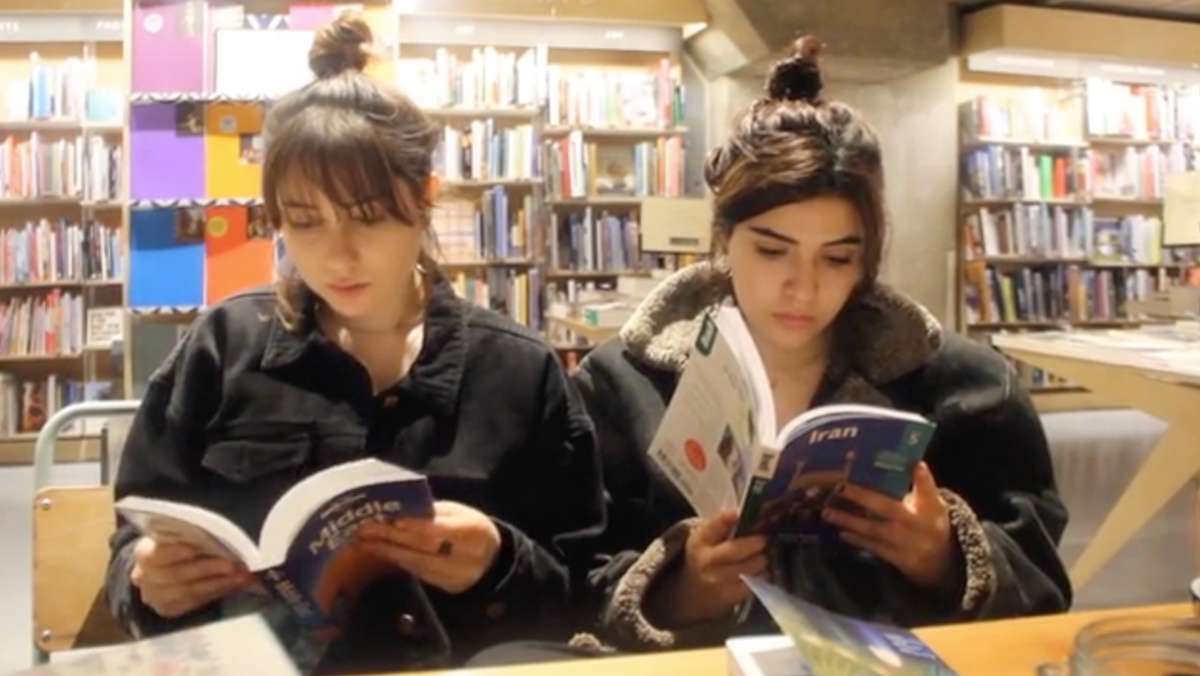 Two young women stand next to each other in a bookshop. They are each reading a travel book.