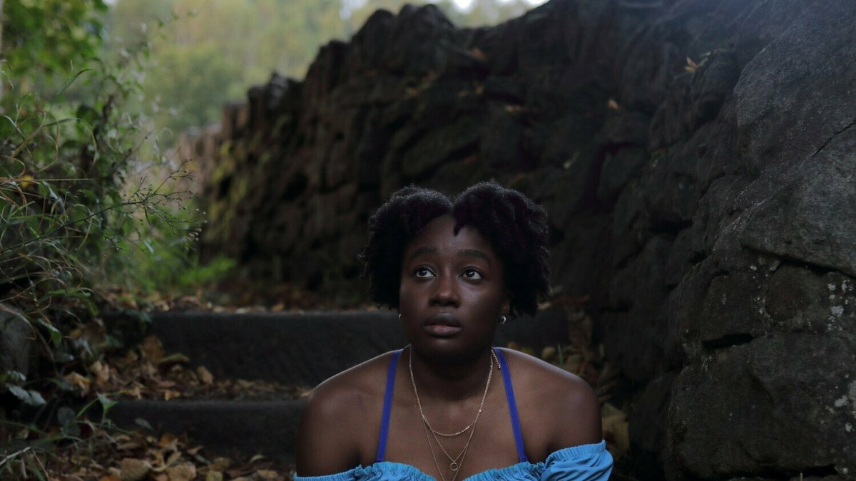 A young black girl in a blue dress sits on the stoops in a forest. She is looking up at the sky.