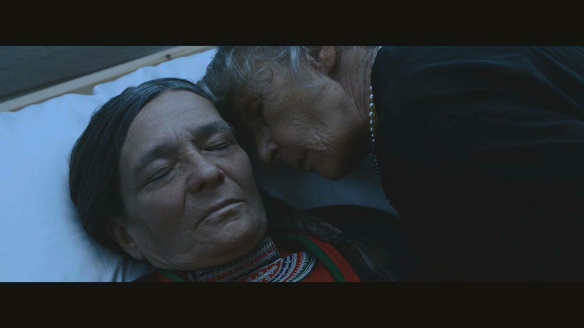 An older woman rests her face on another mature woman laying in a coffin.