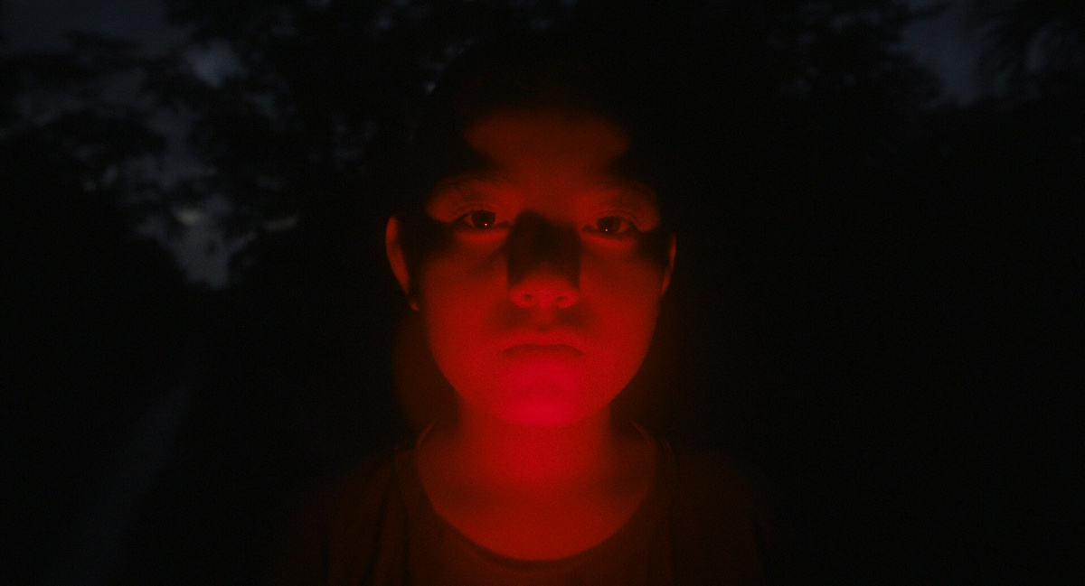 The face of an Indigenous Mexican boy is illuminated from below by a red light.