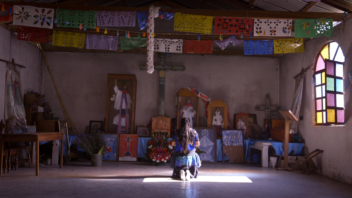An Indigenous Mexican woman in traditional dress kneels inside a church, in front of religious paintings and statues