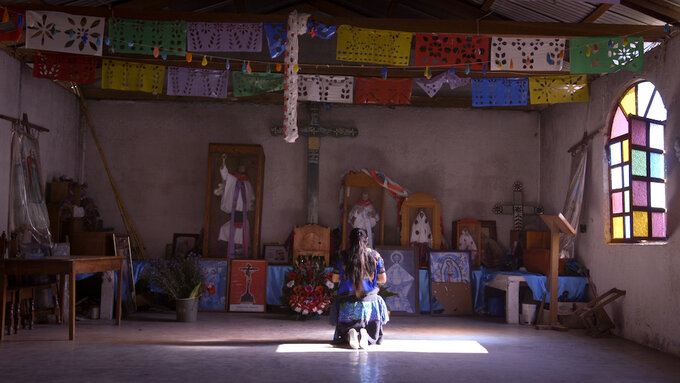 An Indigenous Mexican woman in traditional dress kneels inside a church, in front of religious paintings and statues