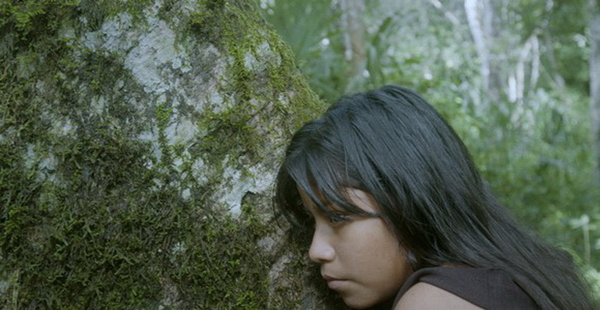 An Indigenous Mexican girl with brown skin and long black hair is leaning her head against a tree trunk in a forest.