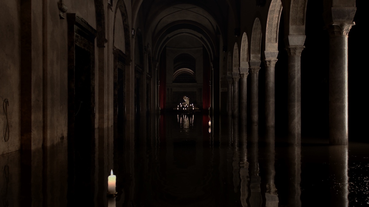 A dark corridor of stone and marble arches, the floor is dark and reflective.