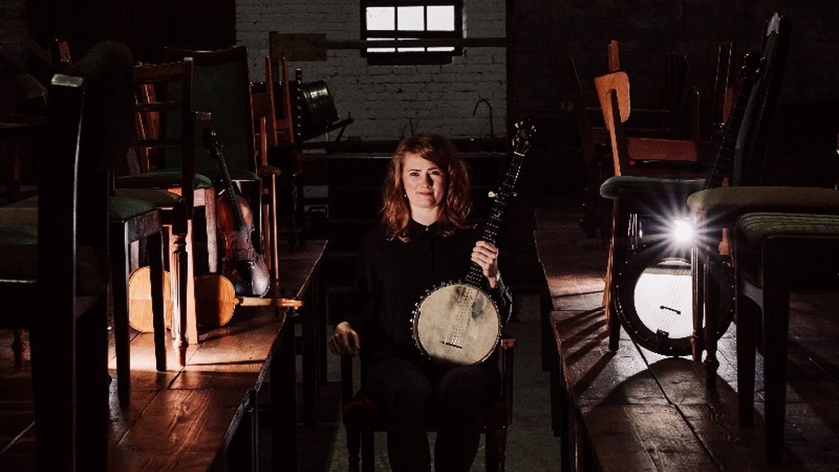 A picture of Rowan Rheingans, she stands holding a banjo in a dark room.
