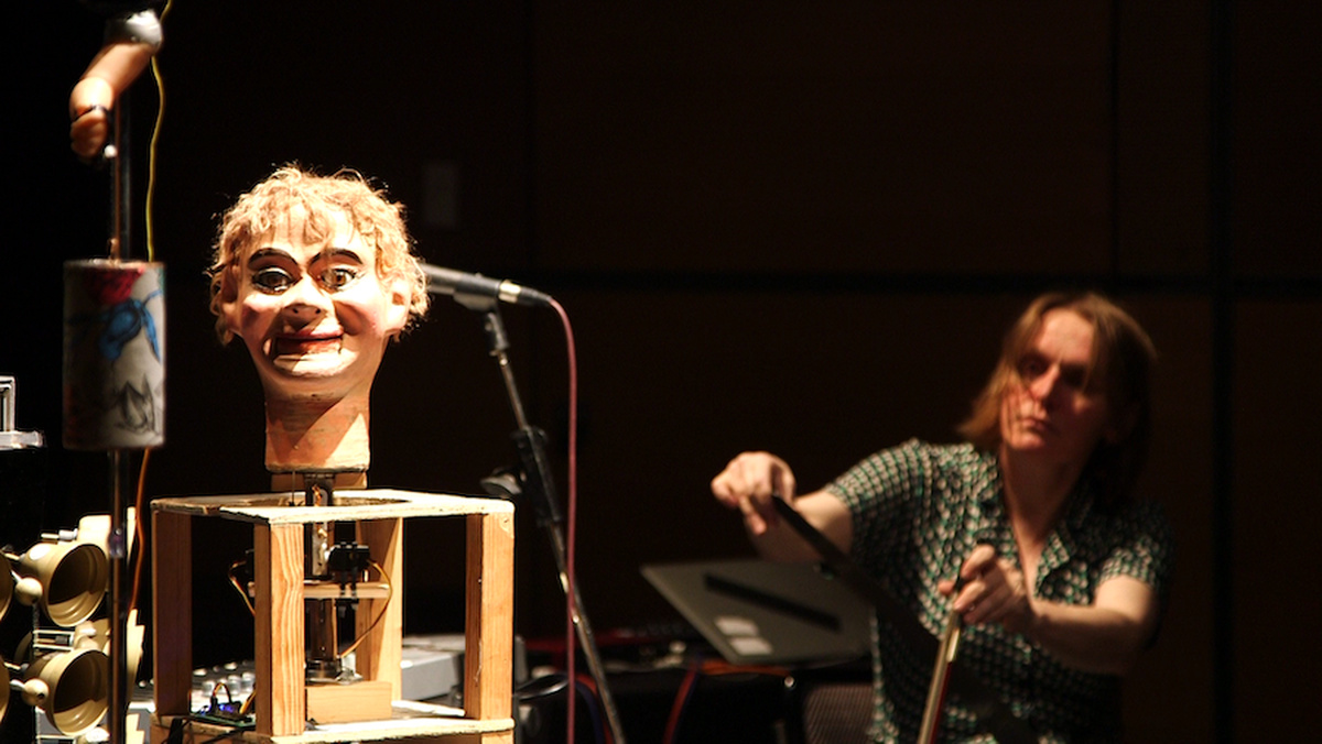 A ventriloquist dummy head and a doll arm mounted on a wooden contraption while a woman plays the theremin.
