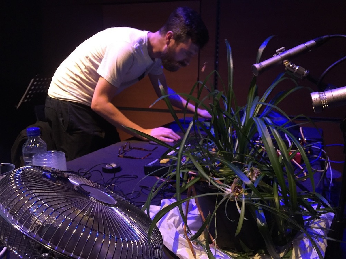A man at a table with mixer, tape loop, electric fan and microphone surrounded by houseplants.