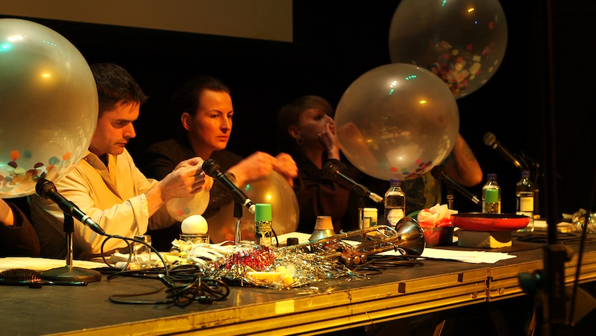 Four people sitting at a table covered with musical instruments and toys are blowing up balloons filled with confetti.