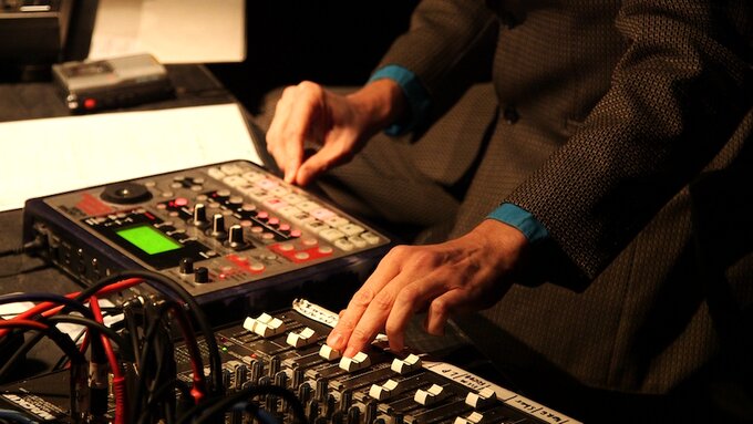 A pair of hands adjusting a mixer on a table covered with electronic equipment and a tape recorder.