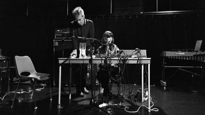 A man is standing and a woman is sitting at an onstage table covered with electronic equipment.