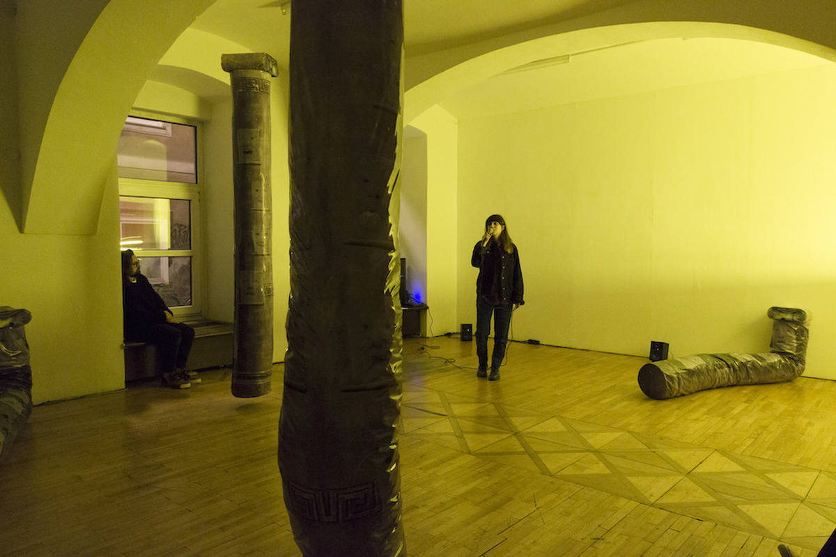 A woman is singing in the middle of a room, surrounded by sculptural tubes and a plith, while a man sits looking on.