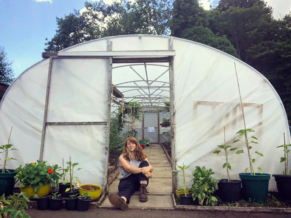 Charley, a person with auburn hair, sits relaxed at the entrance of a polytunnel surrounded by potted plants and flowers