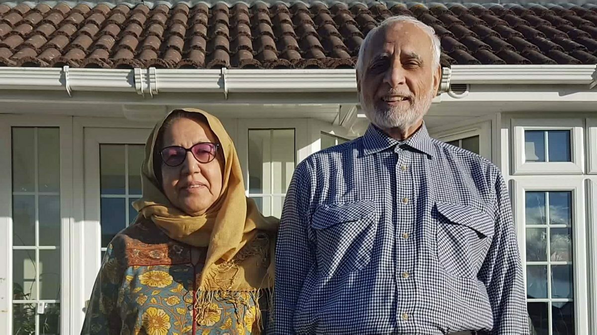 An elderly man and woman from Pakistan are smiling, standing outside their home in Stornoway in the sunshine.