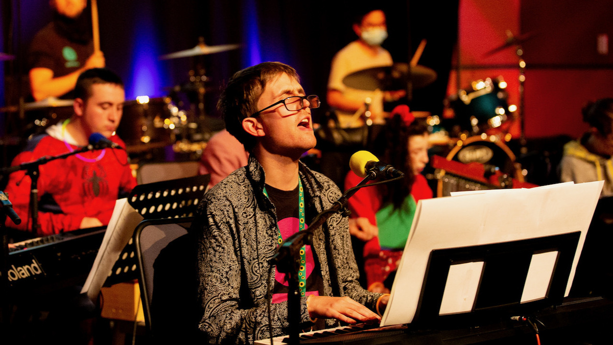 A person with glasses at the keyboard singing into a microphone. With other musicians in the background.