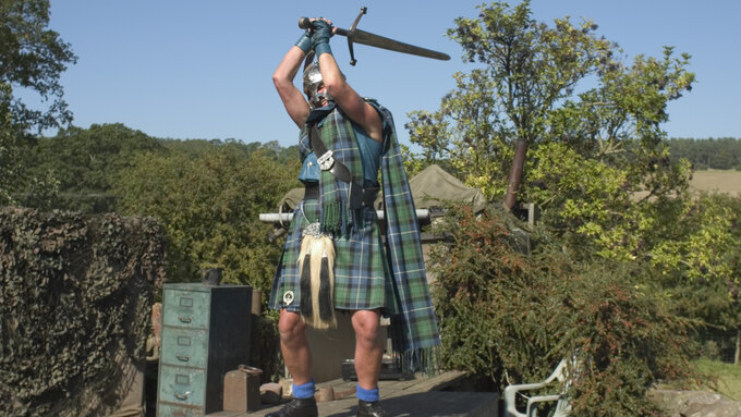 A man wearing traditional Highland dress raises a sword above his head.