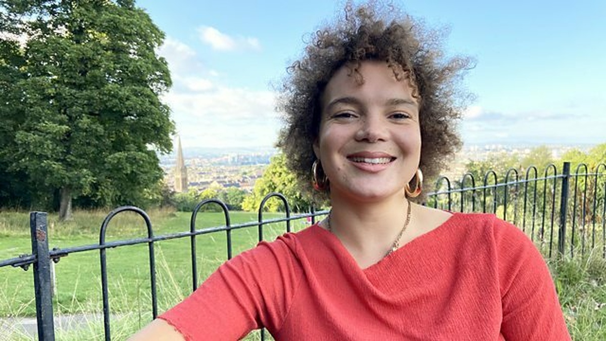 The presenter of the film, Cass Ezeji, is standing in the park, smiling. There is a view overlooking Glasgow behind her.