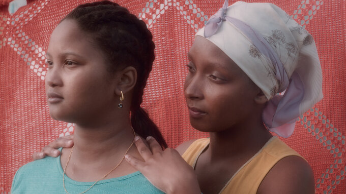 Two Cape Verdean, dark-skinned women stand together. One woman has her hands placed on the other woman's shoulders.