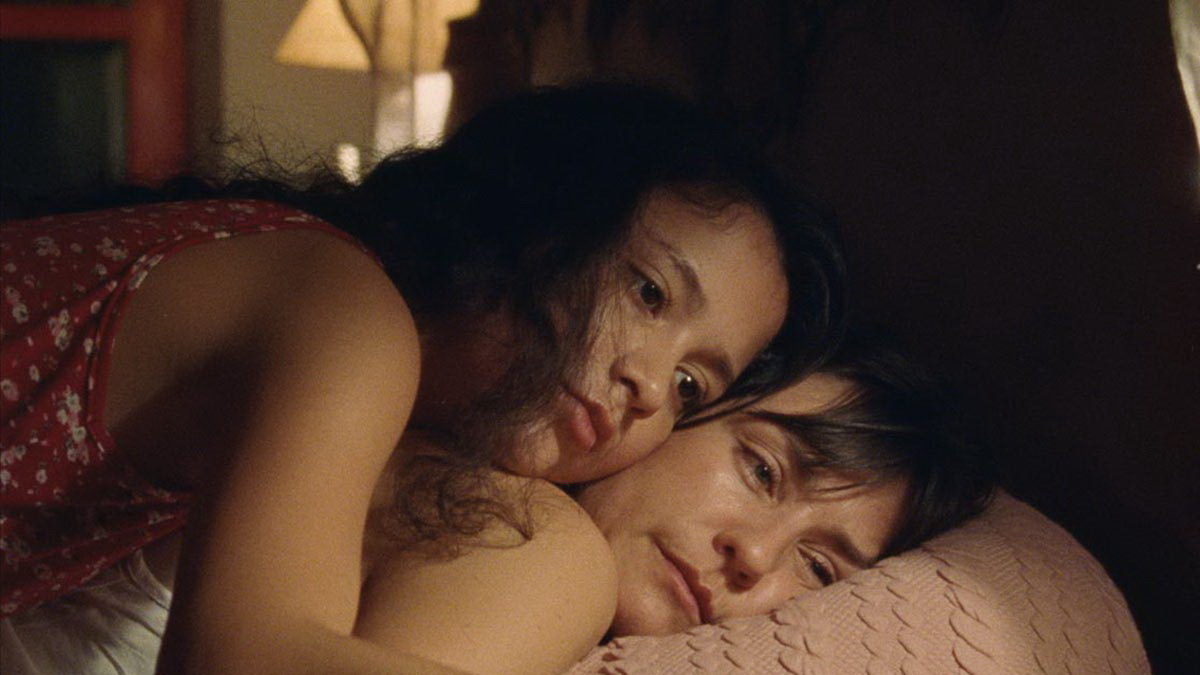 A teenage girl is lying on top of her mother, their cheeks and hands pressed together in tender embrace.