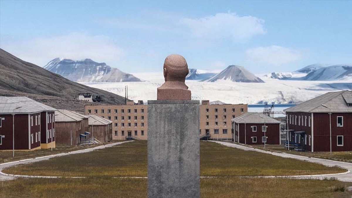 A stone bust sits on a plinth, overlooking a grass pitch, cluster of institutional buildings and snowy mountain range.